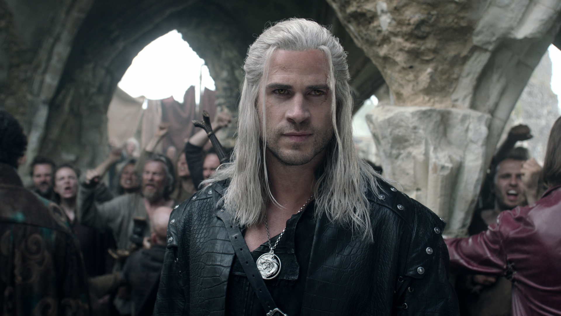 Liam Hemsworth caracterizado como Geralt de Rivia, com cabelos loiros longos, cicatriz no rosto e roupa de couro preta em cenário medieval. Multidão ao fundo celebra.