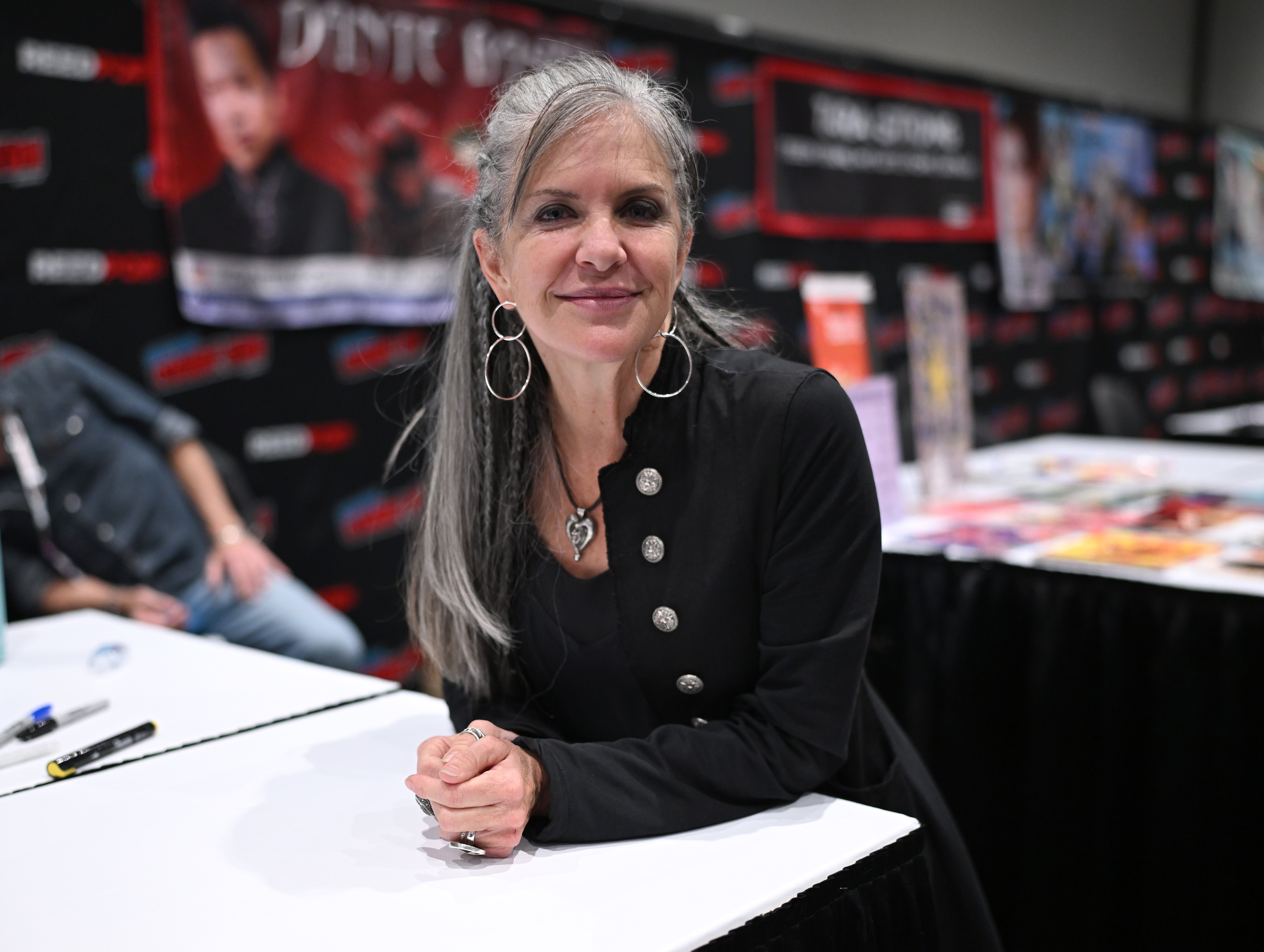 Jennifer Hale, voz da Shepard feminina, revela desejo de voltar ao universo de Mass Effect 1 Jennifer Hale sorrindo, sentada em mesa branca durante convenção, com casaco preto e acessórios prateados