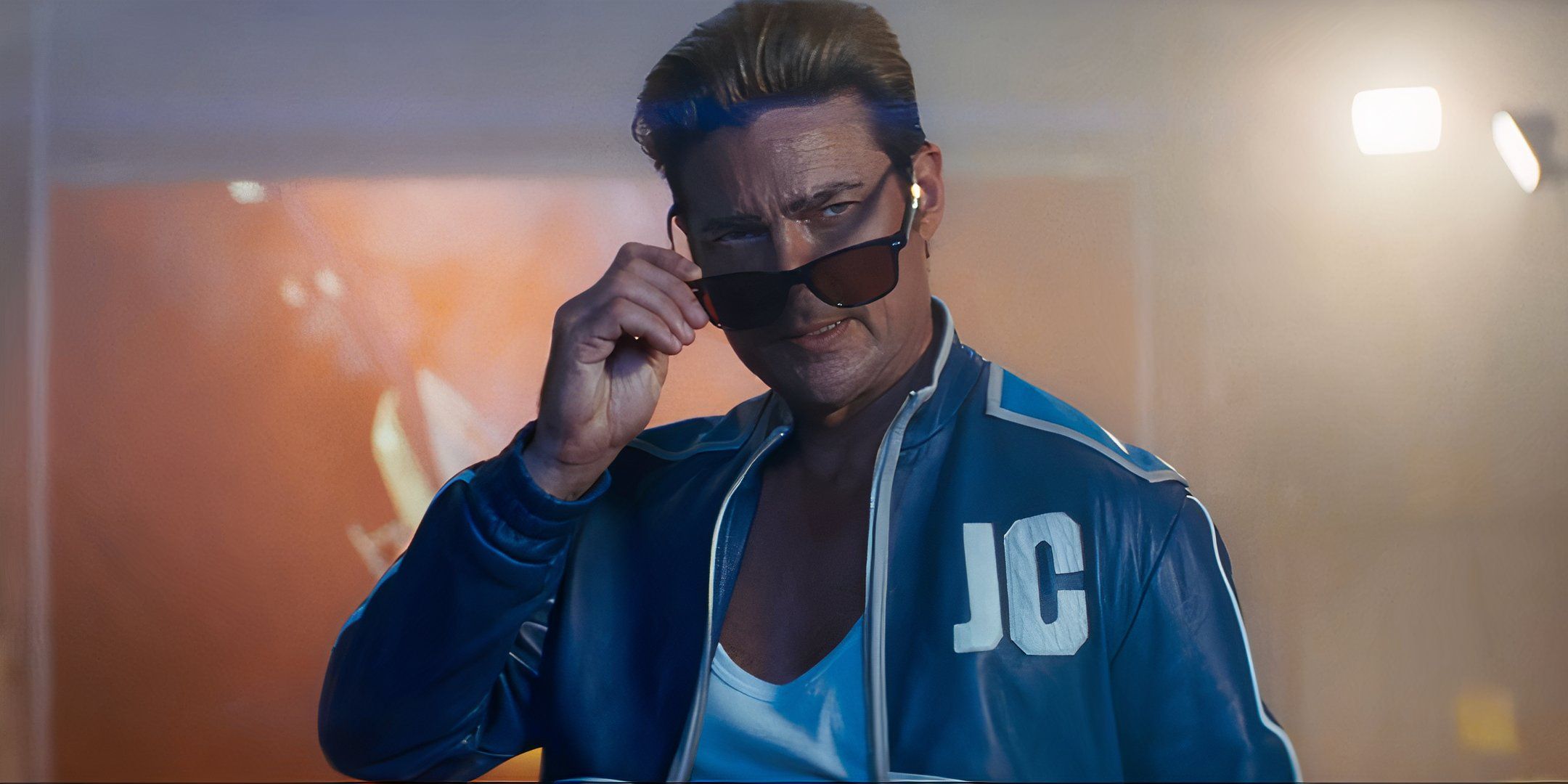Produtor de Mortal Kombat responde provocação do elenco de Street Fighter 1 Personagem Johnny Cage em meio corpo, vestindo jaqueta azul vibrante com as iniciais 'JC', segurando óculos escuros, iluminado em ambiente de estúdio.